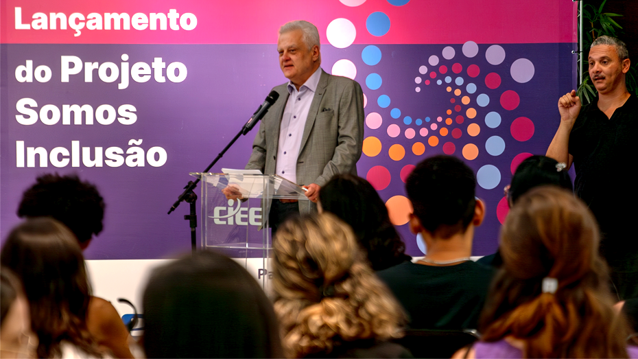 Foto com um banner roxo e rosa escrito “Lançamento do Projeto Somos Inclusão”, a frente, vemos o Humberto Casagrande discursando de frente para uma plateia e ao lado um intérprete de libras.