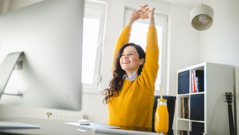 Imagem de uma mulher branca, sentada na frente de seu computador de trabalho se alongando para cuidar da saúde e bem-estar no ambiente de trabalho.